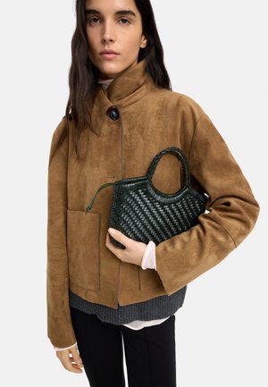 Kobieta w brązowej zamszowej kurtce trzyma małą czarną plecioną torebkę na neutralnym tle.
