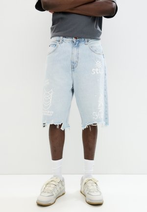 Lyseblå denimshorts med slidte kanter, prydet med hvide grafiske tryk og lapper. Båret med hvide sokker og cremede sneakers.