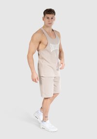Beige sporttanktop met witte tekst, gecombineerd met bijpassende shorts. Gedragen met witte sneakers, wat een ontspannen pasvorm en naadloos ontwerp toont.