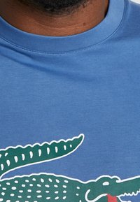 Niebieska bawełniana koszulka z grafiką zielonego aligatora z białymi plamkami i czerwonym językiem, okrągłym dekoltem i krótkimi rękawami.