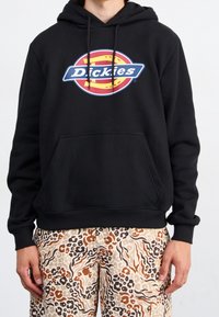 Osoba v černé mikině s barevným logem Dickies a světle béžovými, hnědými a černými kalhotami se zvířecím potiskem stojí proti bílému pozadí.