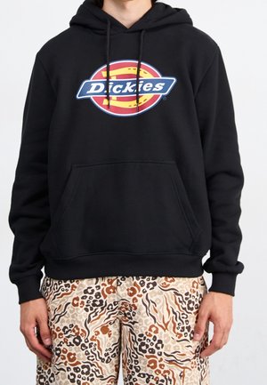 Persona che indossa un hoodie nero con un logo Dickies colorato e pantaloni a stampa animale beige chiaro, marrone e neri, in piedi su uno sfondo bianco.