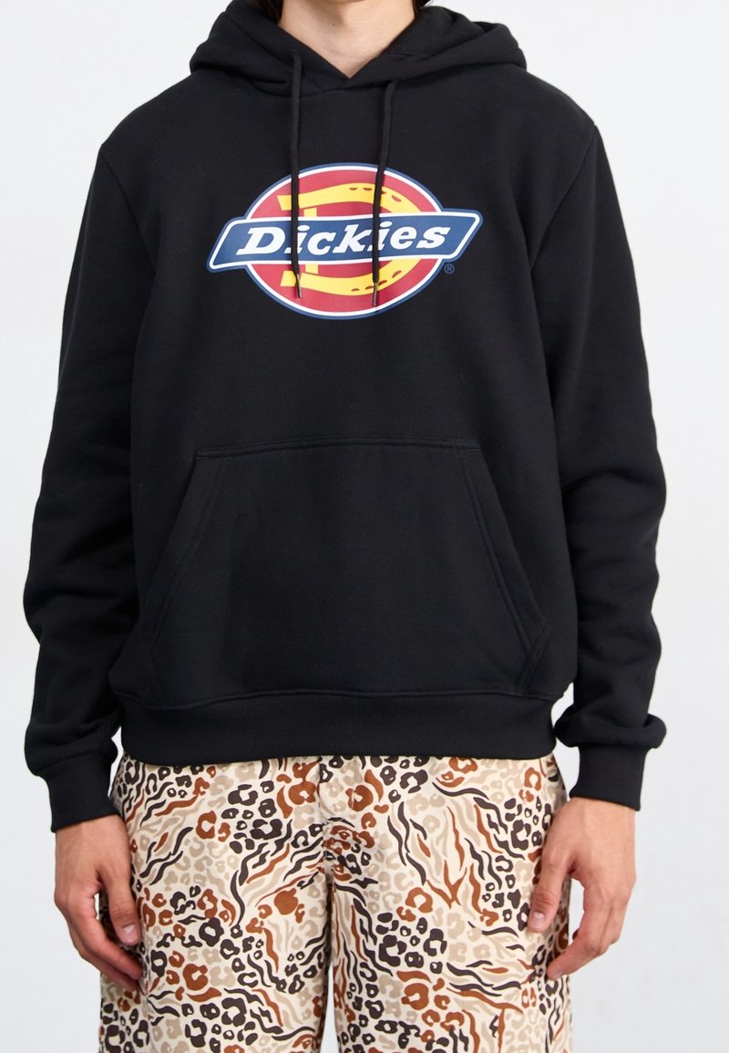 Osoba v černé mikině s barevným logem Dickies a světle béžovými, hnědými a černými kalhotami se zvířecím potiskem stojí proti bílému pozadí.