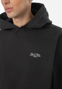 Hanorac negru cu glugă, având un logo frontal din text alb, fabricat dintr-un material moale cu o textură netedă și un design casual.
