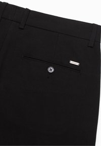 Svarta skräddarsydda byxor med en bakficka som knäpps med knapp och en liten metallmärkning ovanför fickan på högra sidan.