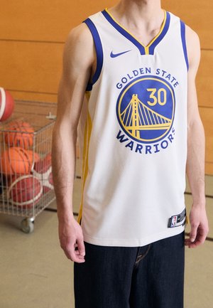 Osoba ubrana w białą koszulkę do koszykówki Golden State Warriors z numerem 30, stojąca obok wózka wypełnionego piłkami do koszykówki.