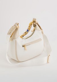 Bílá látková taška přes rameno s béžovými koženými detaily, zlatým zipem, dekorativním zlatým řetízkem s modrými akcenty a nastavitelným širokým popruhem.