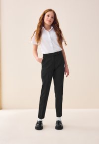Black tailored trousers with a smooth texture, button closure, and front pockets, paired with a white collared shirt. Shoes are glossy black.