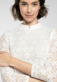 Weißes Spitzentop mit hohem Kragen, floralen Muster und Dreiviertelärmeln. Leichter Stoff mit strukturiertem Überlagerungsdetail.