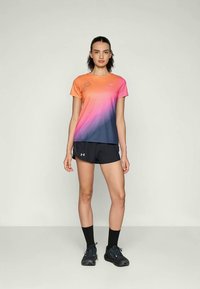 Kvinne står iført en kortermet gradient skjorte i oransje-rosa-blå, svarte shorts, svarte sokker og svarte joggesko, mot en ensfarget bakgrunn.