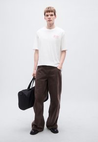 Λευκό βαμβακερό μπλουζάκι με κόκκινο λογότυπο, καφέ παντελόνι φαρδιάς γραμμής από υφασμάτινη υφή, μαύρη τσάντα duffel με κυκλική βάση και μαύρα παπούτσια.