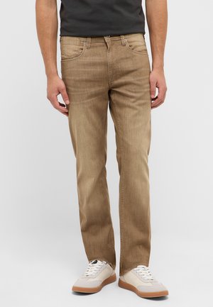 Man wearing light brown straight-fit jeans and white sneakers, standing against a plain light gray background.