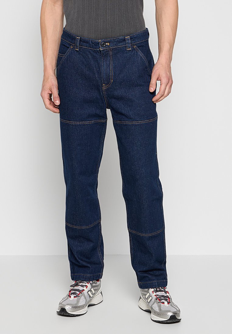 Donkerblauwe denim jeans met een standaard pasvorm, voorzien van contrasterende gele stiksels en voorzakken, gedragen met grijze sportschoenen.