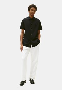 Camicia nera a maniche corte con colletto button-down, caratterizzata da un logo ricamato, abbinata a pantaloni bianchi e scarpe nere.