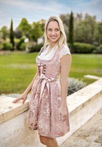 Traditionelles pinkes Dirndl mit Spitzenärmeln, floralem Stickmuster und einer Satinschleife in der Taille. Vor einem grünen Gartenhintergrund.