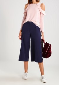 Top spalle scoperte di un rosa chiaro con dettagli arricciati, abbinato a culotte a gamba larga blu navy e sneakers bianche. Accessorio zaino color bordeaux.