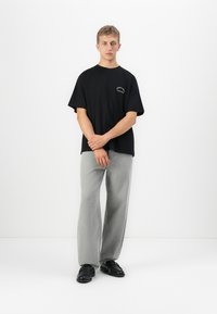 Ung mand står op ad en hvid væg iført en sort oversized T-shirt, lysegrå vide bukser og sorte lædersko.