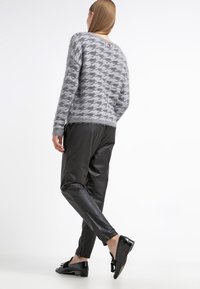 Donna con capelli lunghi che indossa un maglione grigio in stile gessato, pantaloni di pelle nera e mocassini neri con frange, in piedi davanti a uno sfondo bianco.