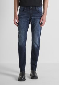 Tamno plavi traperici sa slim fit krojem, s laganim izblijedjelim efektom, pet džepova i metalnim zakačenjem, u kombinaciji s crnim čizmama.