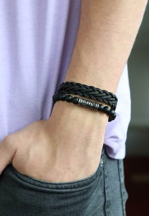 Zwarte gevlochten leren armband en zwarte kralenarmband met een zilveren decoratief element, gedragen om een pols met een lichte huidskleur.