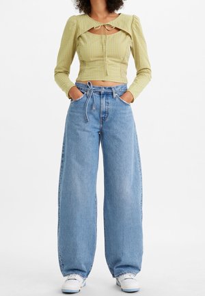 Frau trägt ein hellgrünes Crop-Top mit Frontknoten und Ausschnitt, kombiniert mit hoch taillierten, weit geschnittenen blauen Jeans und weißen Sneakers, Hände in den Taschen.