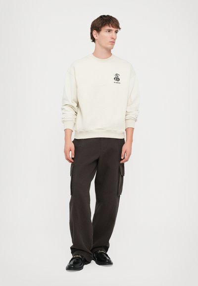 Lys cremefarvet sweatshirt med logo og ribbede manchetter, parret med mørkebrune cargo bukser med sidelommer, båret med sorte sko.