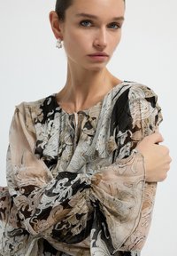 Blusa trasparente con intricate fantasie paisley nere, crema e oro. Maniche arricciate, scollatura con lacci e design fluido.