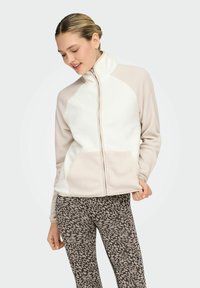 Chaqueta de forro polar en crema y beige claro con cuello alto, cremallera completa y dos bolsillos frontales. Combinada con leggings marrones con estampado.