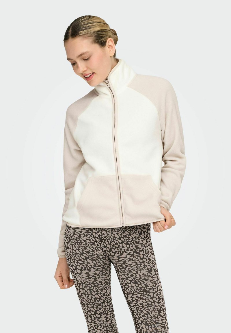 Chaqueta de forro polar en crema y beige claro con cuello alto, cremallera completa y dos bolsillos frontales. Combinada con leggings marrones con estampado.