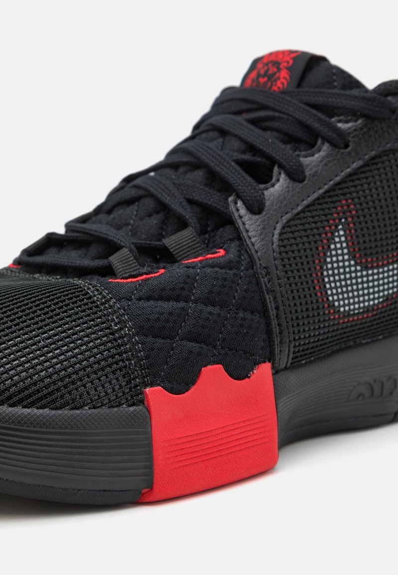 Chaussure de sport noire avec des textures en maille et matelassées. Présente des accents rouges, une semelle en caoutchouc et un logo swoosh bien visible sur le côté.
