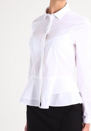 Biała bluzka z długim rękawem, z warstwowym dołem w stylu peplum, zapięta na guziki z przodu, z ostrym kołnierzem, noszona z czarną spódnicą lub spodniami.