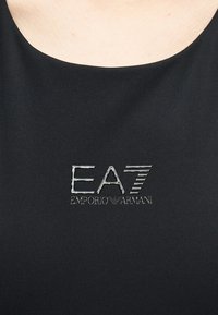 Κοντινό πλάνο μαύρου υφάσματος με το ασημένιο λογότυπο "EA7 Emporio Armani" τοποθετημένο στο κέντρο κάτω από το λαιμό.