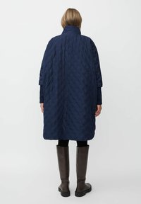 Abrigo acolchado azul marino con un patrón texturizado, diseño oversize, cuello alto y mangas de tres cuartos, combinado con botas marrones hasta la rodilla.