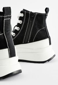 Sneakers alte nere con cuciture bianche, suola bianca spessa e cerniera laterale. Tessuto con dettagli stringati e suola in gomma nera.