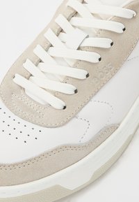 Sneaker in pelle bianca con dettagli in suede beige. Presenta lacci bianchi piatti, punta perforata e suola in gomma testurizzata. Logo "BOSS" visibile.
