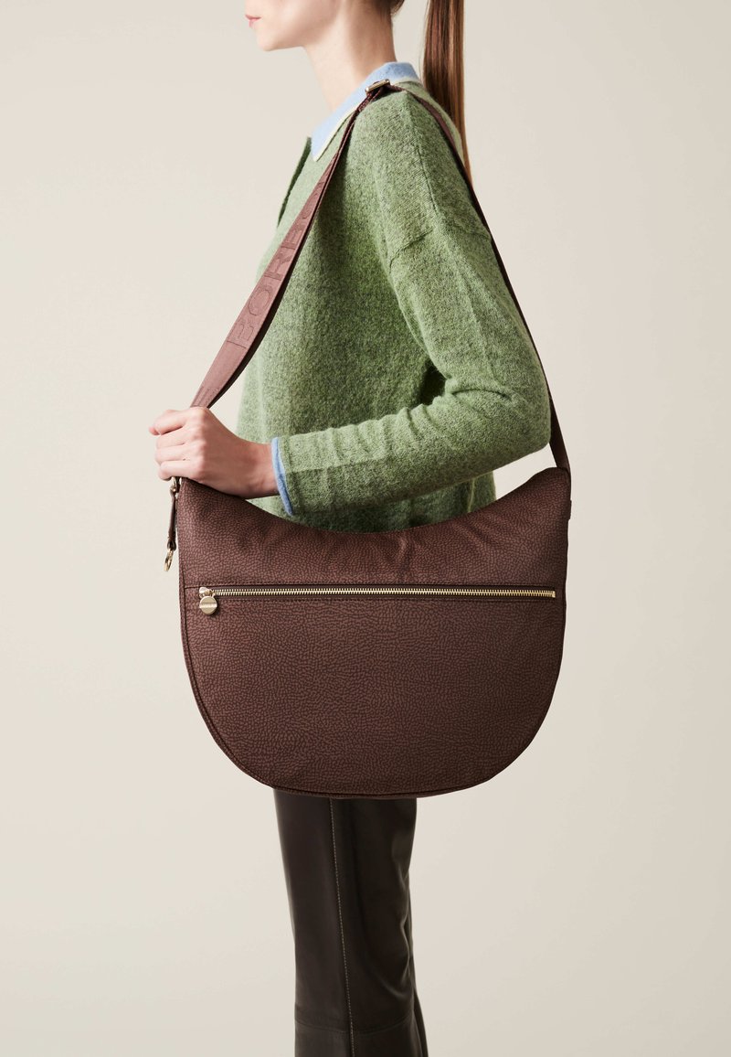 Braune strukturierte Schultertasche mit geschwungener Form und goldenen Reißverschlussdetails, crossbody getragen von einer Person in einem grünen Pullover.