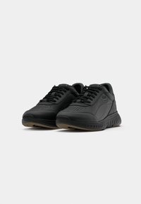 Zapatillas deportivas negras de cuero, con un diseño perforado, lengüeta texturizada y suela flexible con acentos marrones en la parte inferior.