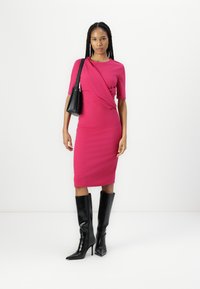 Robe ajustée fuchsia à manches courtes, silhouette ajustée et détail drapé, associée à des bottes noires en cuir verni à hauteur de genoux.