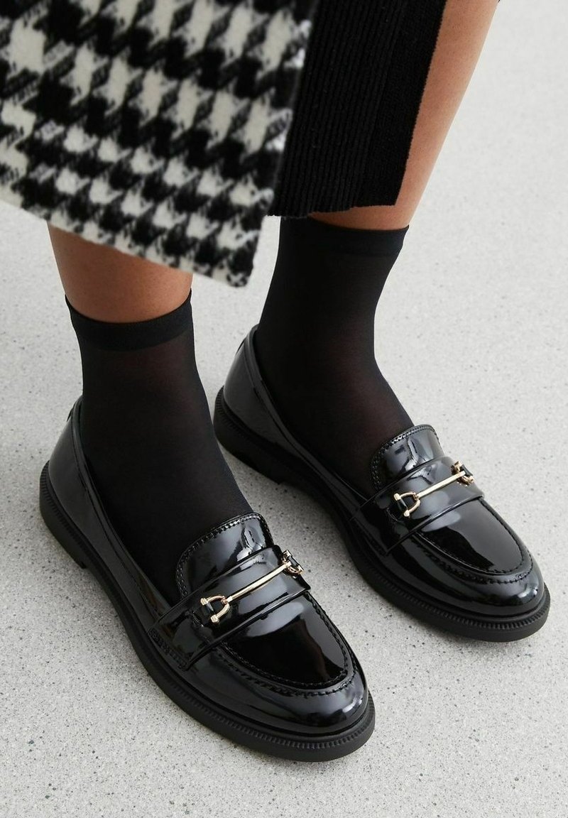 Mocassins en cuir verni noir à finition lisse, ornés d'accents en métal doré et d'une semelle texturée. Portés avec des chaussettes noires transparentes.