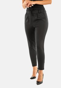 Pantalones ajustados negros con un patrón texturizado, cintura con cordón, y dos bolsillos laterales, combinados con tacones altos negros de charol.