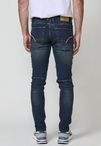Jeans ajustados de color azul oscuro con un acabado desgastado, que presentan detalles de costura amarilla y bolsillos traseros. Combinados con zapatillas grises y azules.