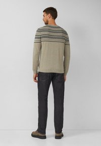 Man in een beige gebreide trui, zwarte jeans en bruine laarzen die voor een effen lichtgrijze muur staat.