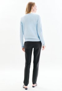 Donna con lunghi capelli biondi che indossa un maglione azzurro chiaro, pantaloni neri e ballerine nere aperte sul retro, in piedi davanti a uno sfondo bianco luminoso.