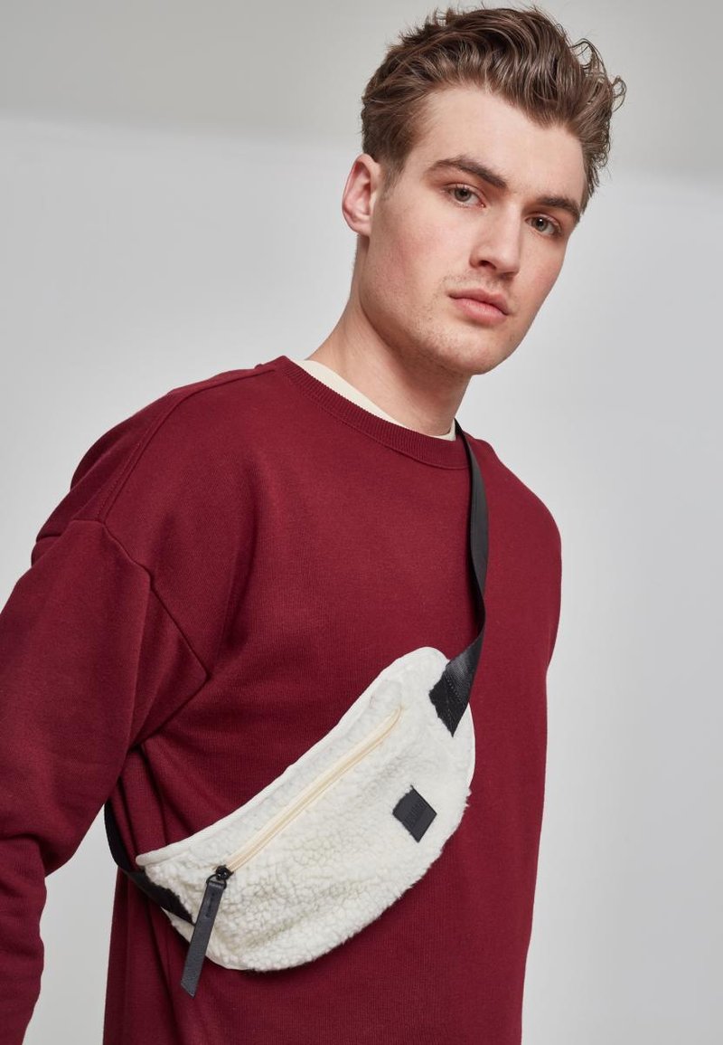 Fleece belteveske i off-white, med en kontrasterende svart glidelås og stropp. Den avrundede formen inkluderer en liten, svart detaljlabel.