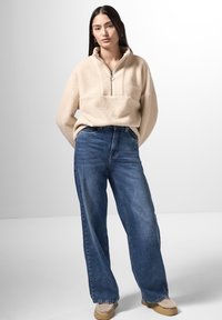 Pullover in fleece beige con colletto con zip, jeans blu a gamba larga e scarpe mocassino color beige. Il modello è in piedi contro uno sfondo neutro.