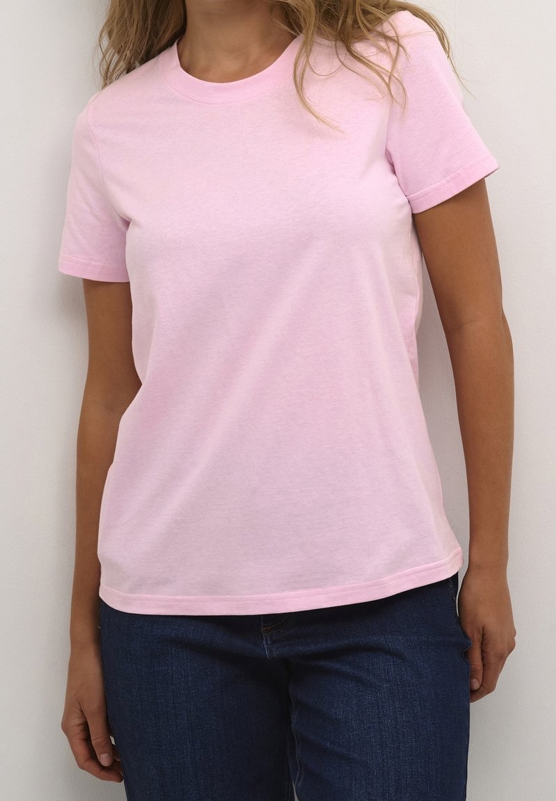 Femme portant un t-shirt rose clair uni à manches courtes et un jean bleu foncé, debout devant un fond blanc.