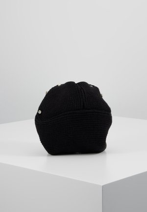 Bonnet en tricot noir avec de petites perles décoratives brillantes placées à la verticale sur une surface blanche en coin, sur un fond gris.