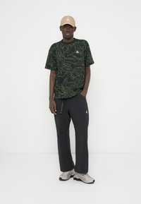 Μαύρο μπλουζάκι με πράσινο σχέδιο; μαύρο παντελόνι φαρδιάς εφαρμογής; μπεζ καπέλο; γκρι αθλητικά παπούτσια με υφή σχεδίου; ορατά λογότυπα.