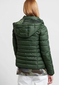 Grön puffjacka med kviltat mönster, huva och dragkedja. Materialet har en glänsande yta, och en kamouflage t-shirt under.