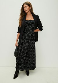Black polka dot maxi dress with a square neckline, layered design, and short black blazer. Paired with glossy black pointed boots.